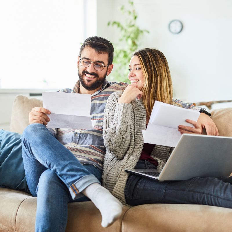 A man and a woman sit on a couch in their home, looking through paperwork. The woman is looking over her shoulder at her partner while balancing a laptop on her lap.