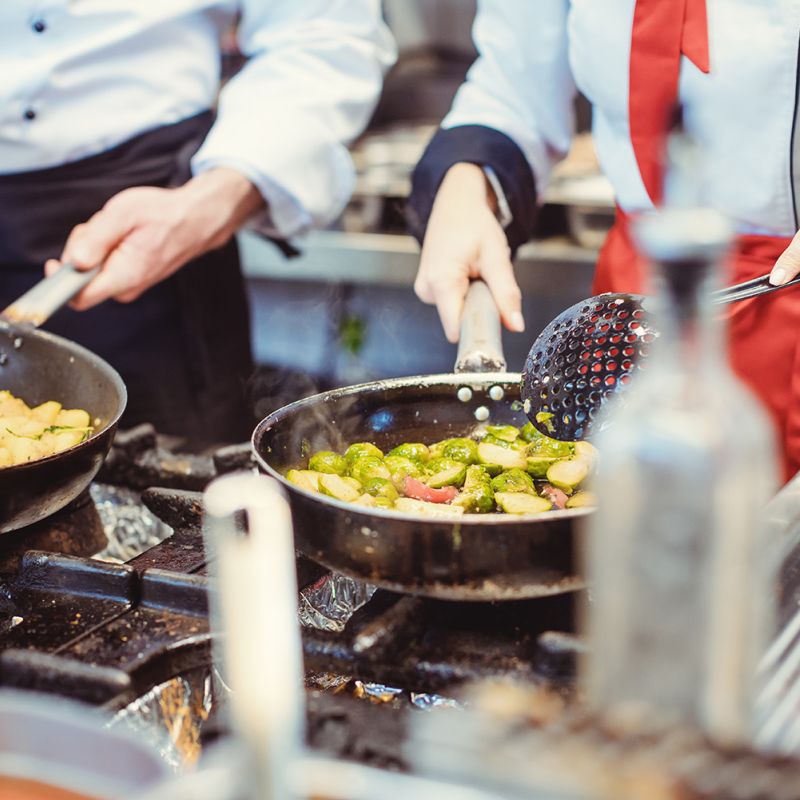 Two people cooking food in woks over gas cookers 
