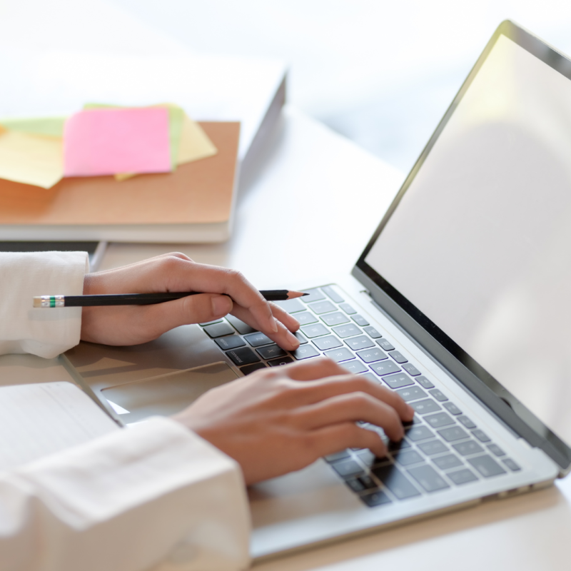 Close up of a person's hands typing on a laptop, with a notebook and post-it notes in the background.
