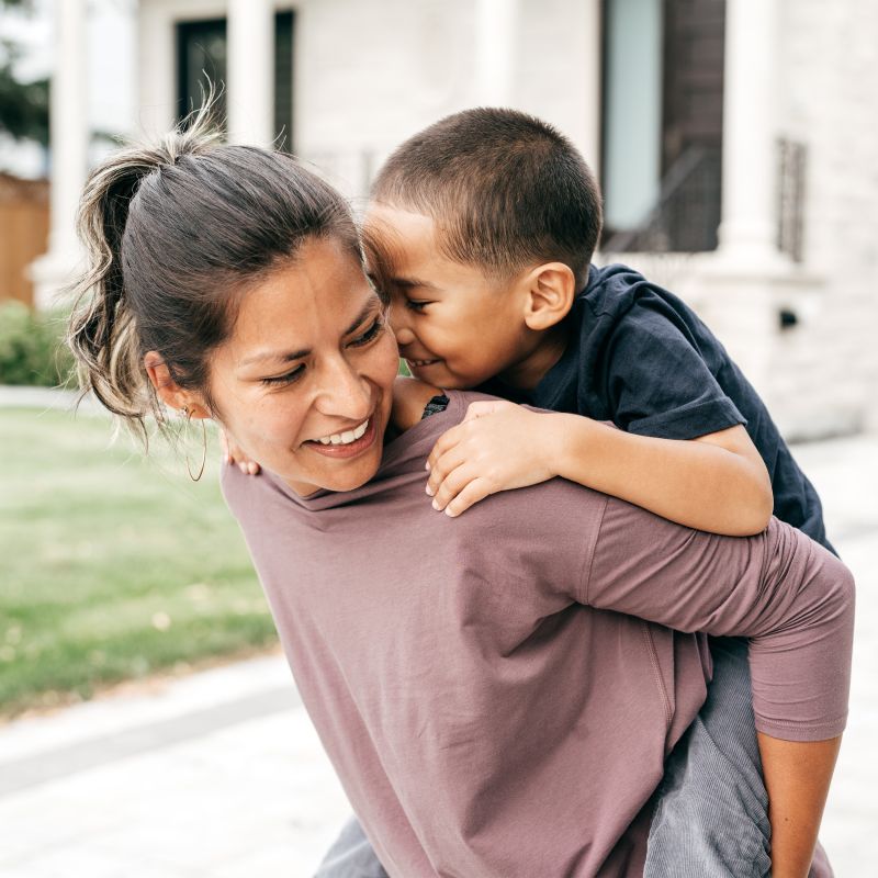 A smiling woman looks over her shoulder as she gives her young son a piggy back ride in the backyard.