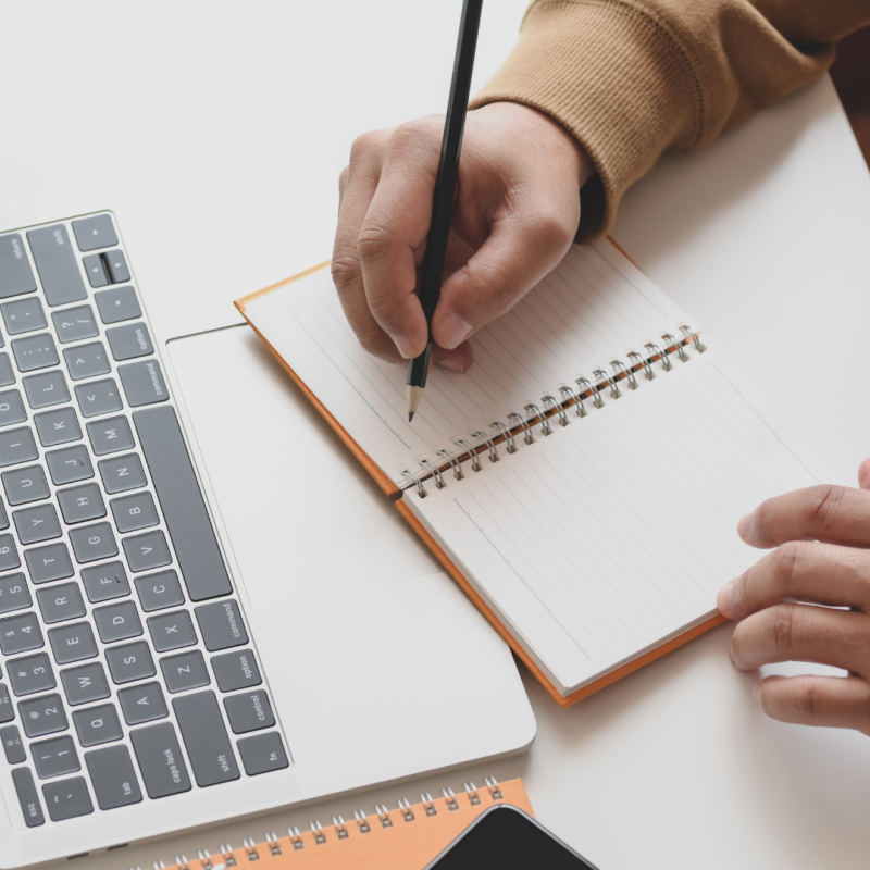 Close up of person writing in a notebook with a laptop in front of them.