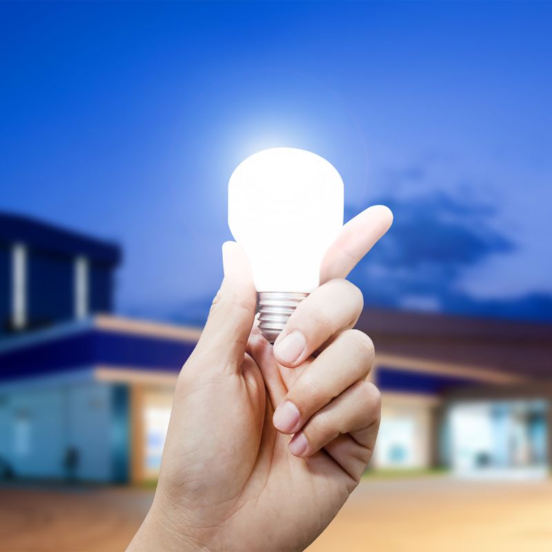 A close up of a person's hand holding a glowing light bulb, in front of a modern house.