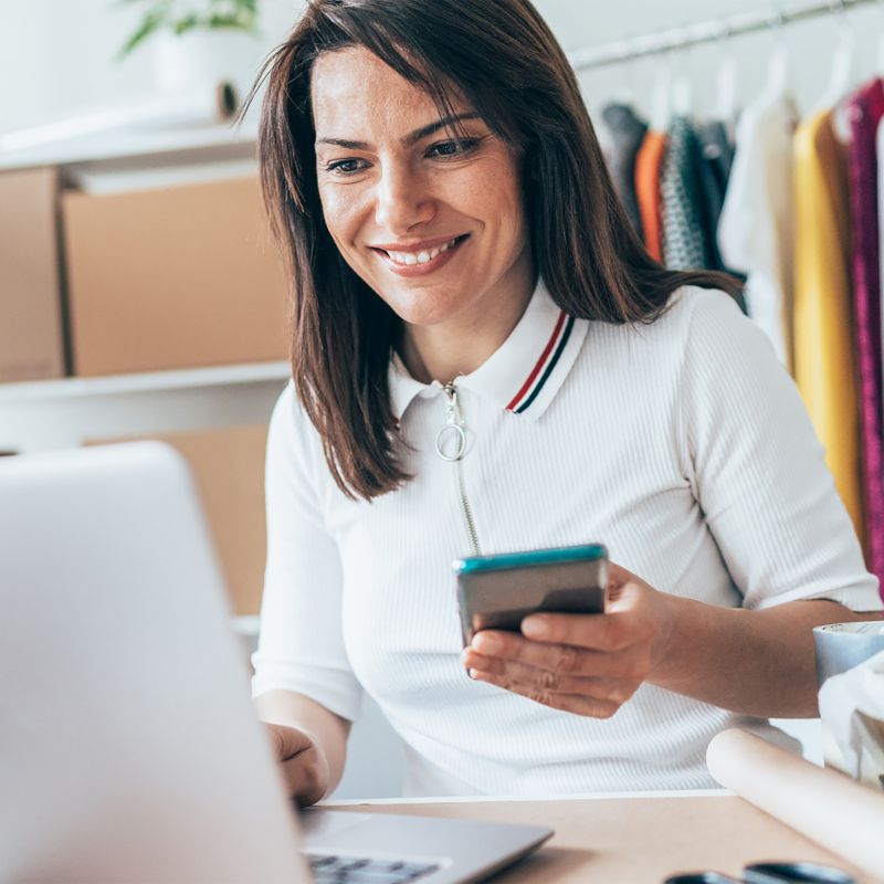 Small business owner looking at her computer with her phone in her hand