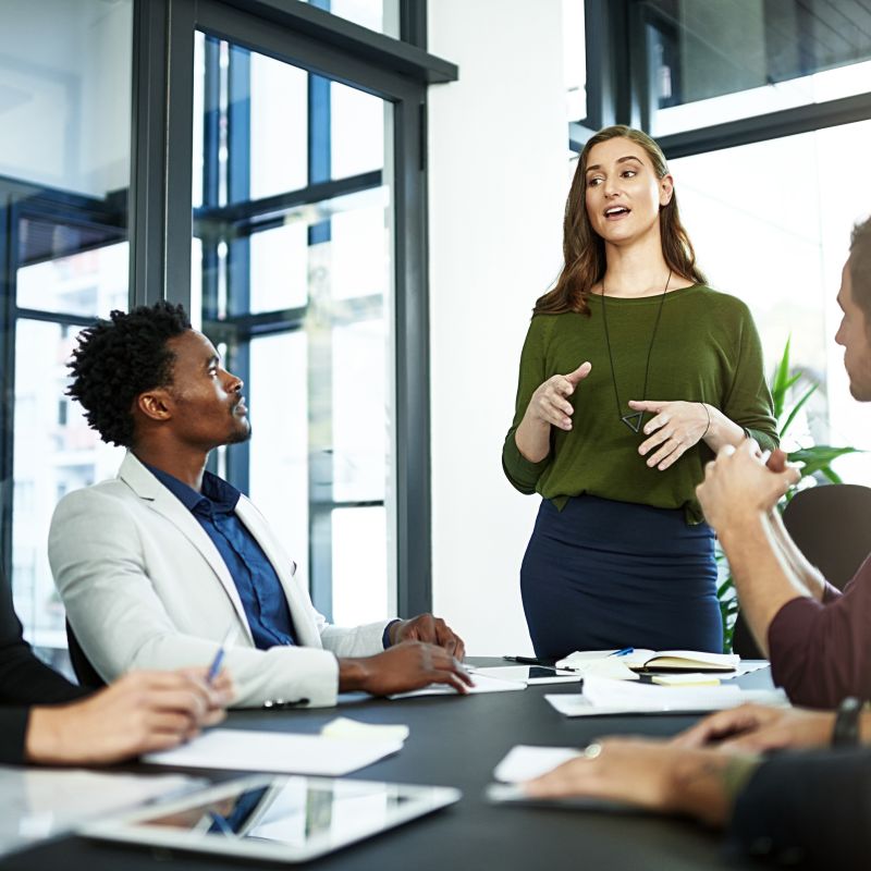 Woman in a green top stands at the head of a boardroom table, presenting to four seated colleagues. 
