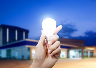 A close up of a person's hand holding a glowing light bulb, in front of a modern house.