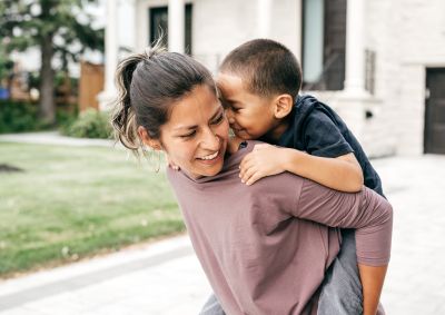A photo of a woman looking over her shoulder as she giver her young son a piggy back in the garden.