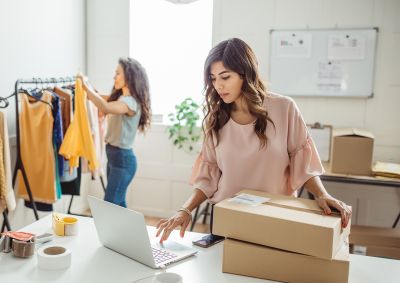 A woman stands in her clothes store processing a transaction on a laptop, with her hand resting on two boxes.