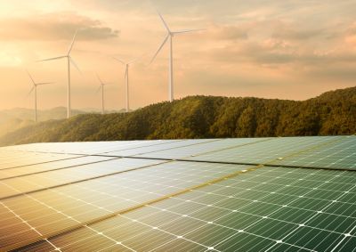 A field of solar panels glistening in the sun with a wind farm on the hills in the background
