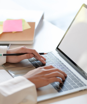 Close up of a person's hands typing on a laptop, with a notebook and post-it notes in the background.