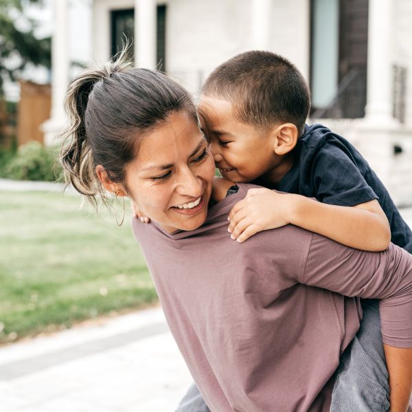 A photo of a woman looking over her shoulder as she giver her young son a piggy back in the garden.